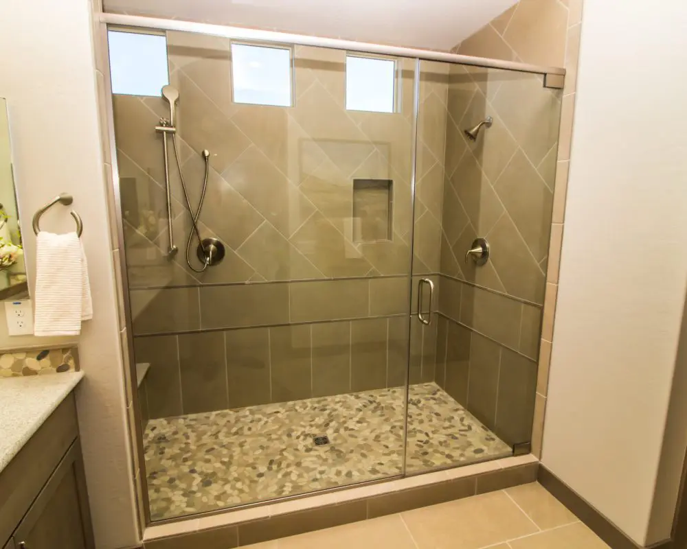 A spacious modern shower with glass doors, three small windows, a handheld showerhead, and pebble-tiled flooring.