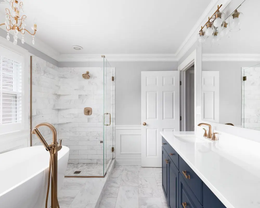 Modern bathroom with a freestanding tub, glass shower, dual sinks on a navy vanity, and brass fixtures. White marble tiles cover the floor and walls. A chandelier hangs from the ceiling.