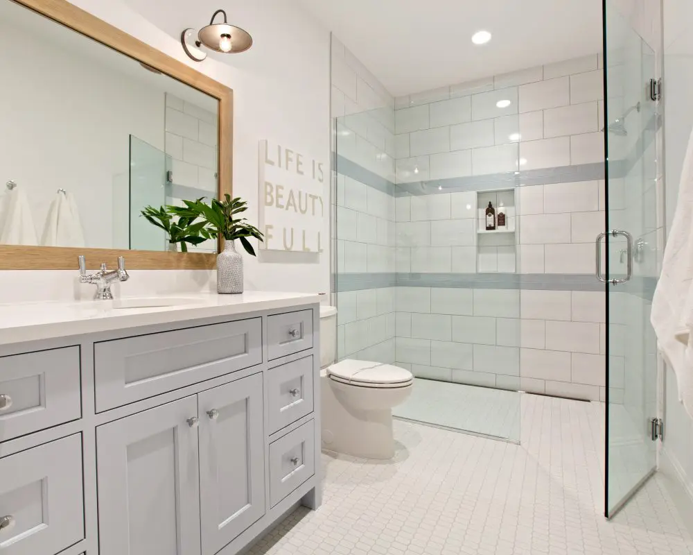 Modern bathroom with a light blue vanity, large mirror, and glass-enclosed shower. A wall sign reads "LIFE IS BEAUTIFUL." White towels hang near greenery on the counter.