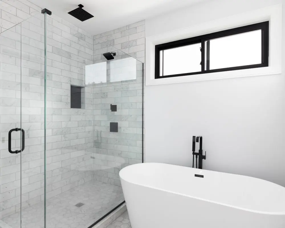 Modern bathroom with a glass-enclosed shower on the left and a freestanding white bathtub on the right. Gray tiles cover the walls, and a black-framed window is above the tub.